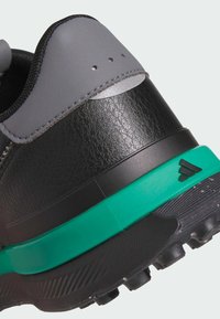 Talons de chaussures noirs et gris en cuir texturé, semelle intermédiaire verte, semelle extérieure noire avec clous, et petit logo noir sur le côté.