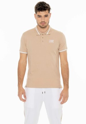 Homme portant un polo beige avec des bordures blanches et un pantalon blanc, debout face à nous sur un fond blanc.