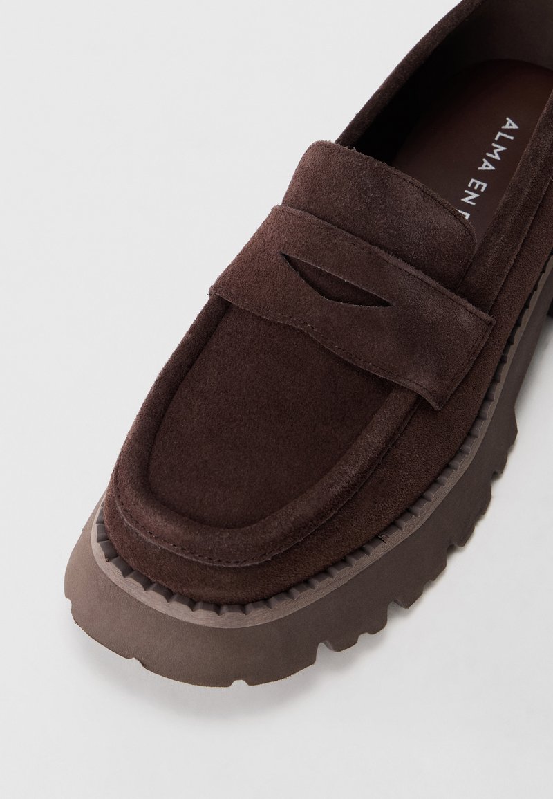 Mocassino in suede marrone scuro con suola spessa a scanalature e dettaglio a fessura sulla fascetta, visto frontalmente su uno sfondo bianco.