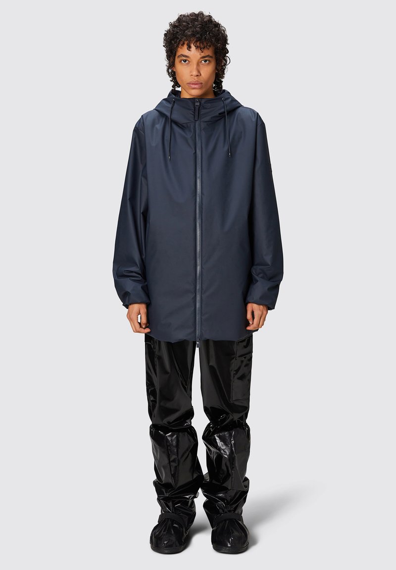 Giacca da pioggia blu navy con cappuccio, chiusura con zip e cordini regolabili, abbinata a pantaloni e scarpe impermeabili neri lucidi.