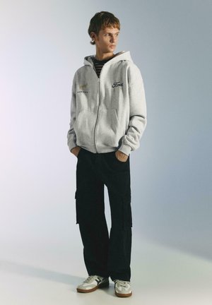 Grå hættetrøje med lynlås og kængurulomme, med broderede logoer og en afslappet pasform. Sammen med sorte cargobukser og hvide sneakers.