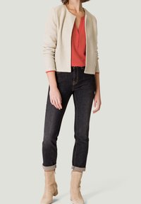 Beige textuur blazer over een rode blouse, gecombineerd met donkere jeans. Model draagt crèmekleurige enkellaarsjes. Blazer heeft geen sluitingen of revers.