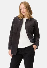 Dunkelbraune Cordjacke mit Knopfverschluss, zwei Brusttaschen und relaxter Passform, getragen über einem weißen Hemd und schwarzen Hosen.