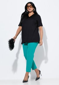 Černý krátký rukávový top s výstřihem do V, spárovaný s modrozelenými formovanými kalhotami a černými vysokými podpatky. Modelka drží černý třásňový doplněk.