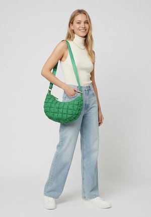 Mujer sonriente que lleva un suéter blanco sin mangas con cuello alto, jeans azul claro, zapatillas blancas y un bolso verde acolchado de hombro con correa larga.