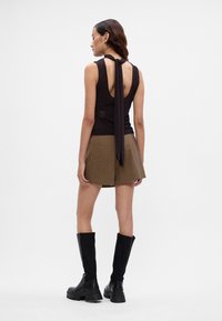 Schwarzes ärmelloses Oberteil mit einer gebundenen Rückenpartie, kombiniert mit beigen karierten Shorts. Das Model trägt knielange schwarze Stiefel.