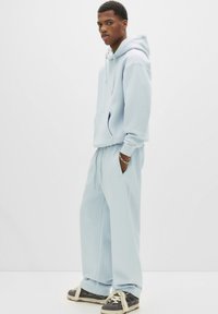 Lichtblauwe hoodie en bijpassende joggingsbroek van zachte stof. De hoodie heeft een kangoeroezak, elastische manchetten en een losse pasvorm. Grijze sneakers.