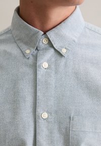 Jasnoniebieska koszula na guziki wykonana z tkaniny tkanej, z ostro zakończonym kołnierzykiem, kieszenią z przodu i białymi plastikowymi guzikami.