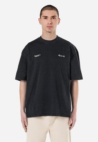 Fekete, oversized pamut póló rövid ujjakkal, bordázott kerek nyakkivágással és fehér hímzett logókkal a mellkason. Lazább és egyszerűbb design.