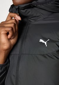 Must tumes, mille kaelusega, sileda tekstuuriga ja valge Puma logoga rinnal. Käsi hoiab tõmbe tõmblukust.