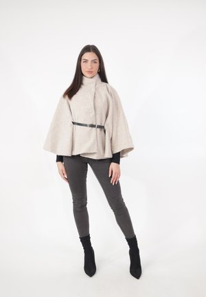 Frau steht und trägt einen beigen, mit Gürtel versehenen Umhang, ein schwarzes langärmeliges Oberteil, dunkelgraue enge Hosen und schwarze spitze Stiefeletten vor weißem Hintergrund.