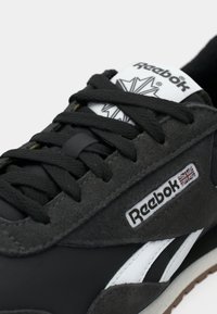 Gros plan sur une basket Reebok noire et grise avec des lacets noirs, un patch logo Reebok blanc et un logo blanc sur la languette.
