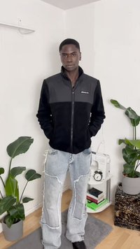 Casaco de fleece preto e cinza com fecho, feito de tecido texturizado, combinado com jeans claros azul desgastados e sapatos pretos. Ambiente interno com plantas.