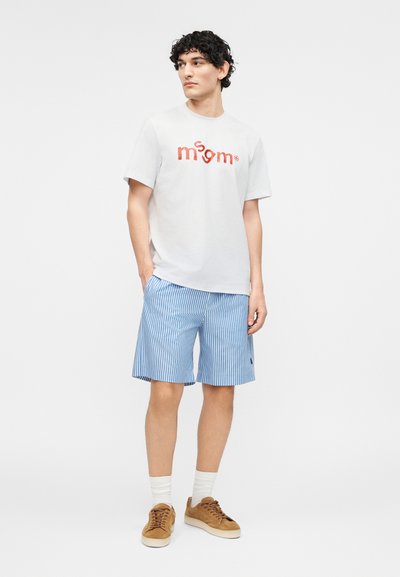 Νέος άνδρας που φοράει λευκό μπλουζάκι με στάμπα, μπλε και λευκές ριγέ βερμούδες, λευκές κάλτσες και καφέ αθλητικά παπούτσια, στέκεται με τα χέρια στις τσέπες.