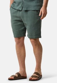 Shorts verts texturés avec une coupe décontractée et des poches latérales, associés à des sandales marron avec deux sangles réglables et une semelle plate.