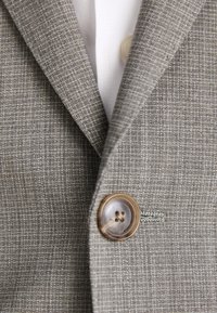 Tessuto di un blazer grigio a scacchi con una trama liscia. In evidenza un pulsante marrone e un colletto di camicia bianco parzialmente visibile.