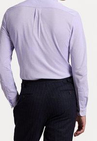 Mężczyzna w jasnofioletowej koszuli z długim rękawem, wsuniętej w ciemne garniturowe spodnie w paski, widziany z tyłu.