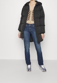 Svart puffjacka med huva, beige stickad cardigan med rutmönster, blå jeans och flerdelade sneakers.