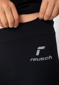 Schwarze Leggings mit einer glatten, dehnbaren Textur und einem hohen Bund. Mit einem silbernen, gestickten "reusch"-Logo an der Seite.