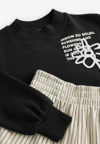 Sweatshirt noir avec col côtelé, orné d'un motif de fleur brodé en blanc et de texte. Associé à un pantalon léger à rayures avec taille élastique.