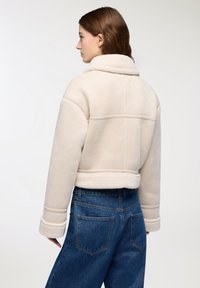 Giacca in shearling beige tagliata con un ampio colletto, finitura testurizzata e maniche con polsini. Abbinata a jeans scuri a vita alta.