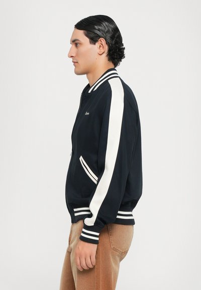 Veste de pilote navy avec des rayures blanches sur les manches, des poignets et une taille côtelés. Fermeture éclair et détail de logo sur la poitrine, associée à un pantalon beige.