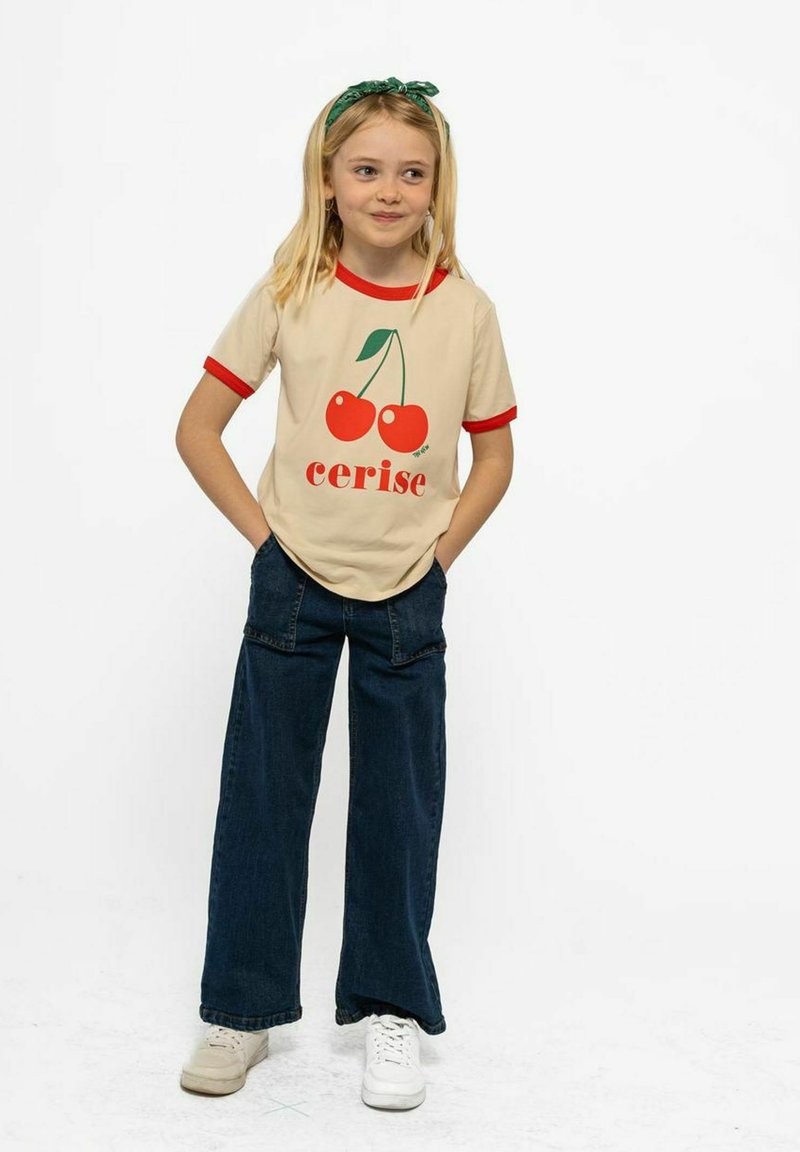 Camiseta beige con ribetes rojos en las mangas que presenta un gráfico de cereza y la palabra "cerise". Combinada con pantalones vaqueros de pierna ancha de color azul y zapatillas.