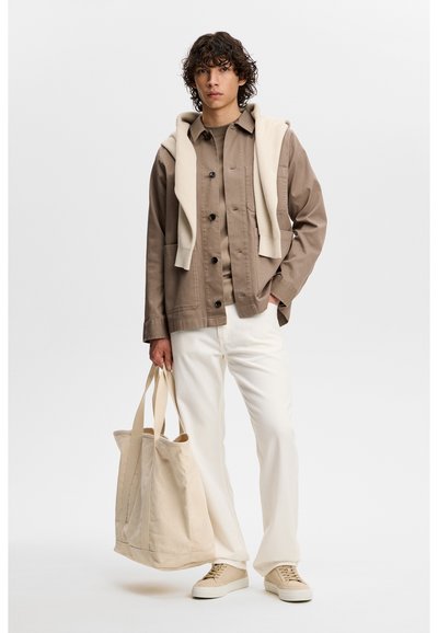 Młody mężczyzna w beżowej kurtce na szarej koszuli, białe spodnie, beżowe trampki, kremowy sweter zarzucony na ramiona, trzymający beżową torbę na ramię.