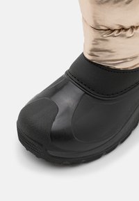 Schwarze wasserdichte Stiefel mit einer strukturierten Gummisohle, ausgestattet mit einem glatten, metallisch gold reflektierenden Obermaterial und einem schwarzen, elastischen Neoprenstulpen.