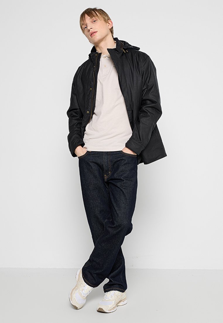 Jeune personne adossée à un mur blanc, portant une veste noire, un polo beige, un jean foncé et des baskets blanches, avec les mains dans les poches.