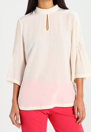 Kvinde iført en cremefarvet bluse med nøglehulshalsudskæring og plisserede trekvartærmer, kombineret med lyserøde bukser.