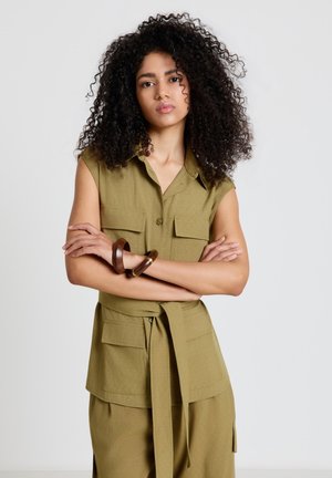 Femme aux cheveux bouclés portant une robe sans manches vert olive boutonnée avec de grandes poches et une ceinture à la taille, les bras croisés et des bracelets en bois.