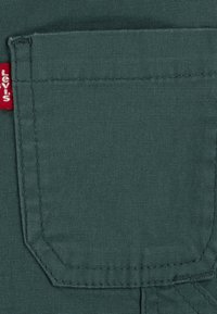 Dunkelgrüner Stoff mit strukturiertem Finish, ausgestattet mit einer Tasche mit abgerundetem Oberteil, verstärkten Nähten und einem roten Logoetikett.