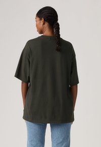 Υπερμεγέθη βαμβακερό μπλουζάκι σκούρου πράσινου χρώματος με κοντά μανίκια, στρογγυλή λαιμόκοψη και ίσια γραμμή, συνδυασμένο με ανοιχτόχρωμα τζιν παντελόνια.