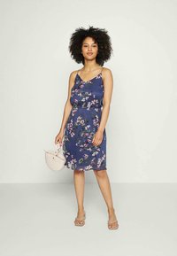 Vestido floral em azul marinho, com um design sem mangas e uma cintura ajustável. A modelo segura uma bolsa beje clara e usa sandálias bejes.