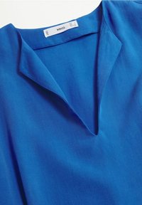 Blouse bleue avec un col en V, une texture douce et un col simple. Le tissu semble léger avec une finition lisse. Étiquette de la marque visible.