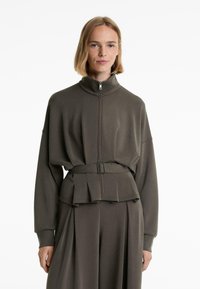 Dark olive green zip-up jacket with a high collar, loose fit, belted waist, and pleated detailing at the hem. Smooth fabric texture.