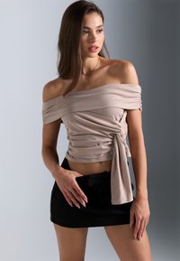 Beige Top mit schulterfreiem Schnitt und gerafften Details, seitlich gebunden, kombiniert mit einem kurzen schwarzen Rock. Glatter Stoff mit eng anliegender Passform.