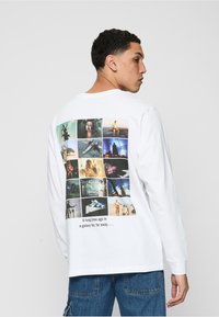 Hvid langærmet T-shirt med et collage af scener fra en sci-fi film på ryggen, med tekst under billederne.