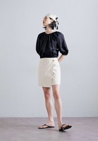 Black puff-sleeve top with elastic neckline, paired with a cream wrap mini skirt featuring a golden button accent and black flip-flops.