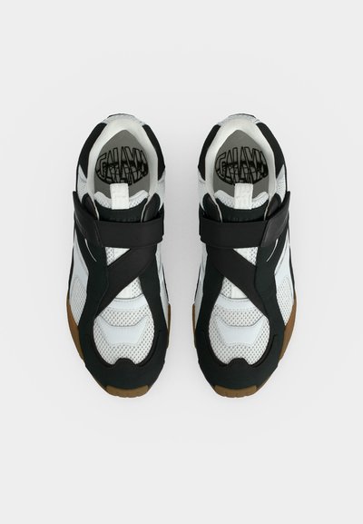 Paire de chaussures de sport blanches et noires avec tissu en mesh, bandes Velcro et semelles en caoutchouc marron vues de dessus.