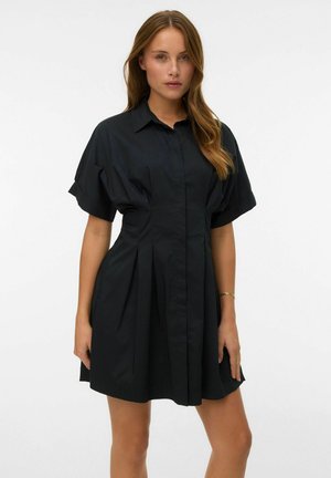 Femme aux longs cheveux portant une robe noire courte boutonnée, manches courtes bouffantes et plis, debout devant un fond blanc.