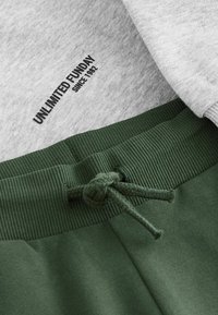 Light gray sweatshirt with "UNLIMITED FUNDAY SINCE 1982" printed on the neckline, paired with dark green joggers featuring a ribbed waistband and drawstring.