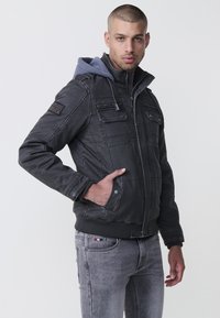 Svart vadderad jacka med grå huva, dragkedjastängning fram och flera bröstfickor. Ribbad mudd och kant, texturerad tyg. Grå jeans synliga.