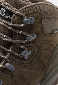 Stivale da trekking marrone realizzato con una combinazione di suede e materiali tessili. Lacci grigi infilati attraverso robusti occhielli con dettagli del logo visibili.