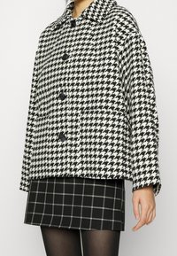Czarny i biały płaszcz w wzór houndstooth z szerokim kołnierzem i czterema guzikami, noszony na czarnej spódnicy w kratkę. Miękka, teksturowana tkanina.