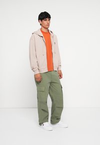 Felpa beige con zip sopra una t-shirt arancione, abbinata a pantaloni cargo verdi. Sneakers bianche con dettagli rossi e blu completano l'outfit.