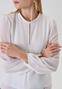 Weiße, durchsichtige Bluse mit Rundhalsausschnitt, Schlüssellochöffnung und puffigen langen Ärmeln. Glatte Textur, minimalistisches Design und leichtes Material.