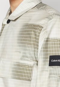 Chaqueta a cuadros translúcida en oliva y blanco, que presenta un cuello, un botón negro, un bolsillo y un detalle del logo de Calvin Klein en el pecho.