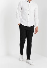 Camicia bianca a maniche lunghe con bottoni neri e tasca, abbinata a pantaloni neri slim-fit e sneakers bianche, in piedi davanti a uno sfondo neutro.